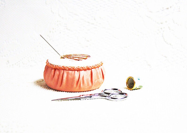 K&ouml;nnte beinhalten: Ein rosa Stoff-N&auml;hkissen mit Nadel und Faden, einer kleinen Garnrolle und einer silbernen Schere auf einer wei&szlig;en Oberfl&auml;che.