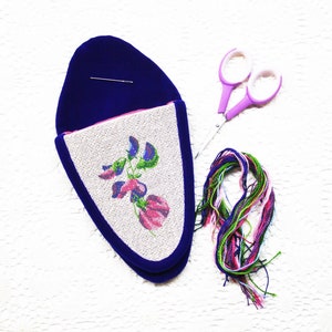 May include: A blue velvet slipper with a white canvas insert featuring a pink and purple embroidered flower design. A pair of purple scissors and a bundle of colorful embroidery floss are also pictured.