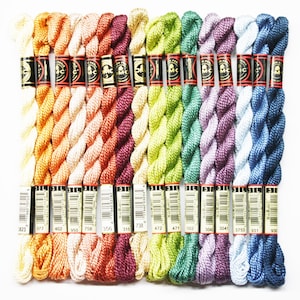 May include: A collection of 15 spools of DMC embroidery floss in various shades of orange, yellow, green, purple, blue, and beige. The spools are labeled with DMC numbers: 1823, 977, 402, 950, 758, 356, 315, 738, 472, 471, 502, 3042, 3041, 3753, 931, and 930.