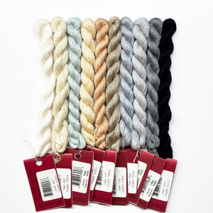 May include: A set of ten skeins of embroidery floss in various shades of gray, beige, and white. The skeins are arranged in a row from darkest to lightest. Each skein has a red label with the color number and dye lot printed on it.