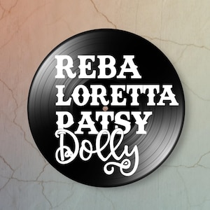 May include: A black circular vinyl record featuring white text printed in the center. The text displays the names REBA, LORETTA, PATSY, and Dolly in a stacked arrangement, with the name Dolly written in a distinct, flowing cursive script font.
