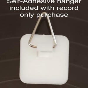K&ouml;nnte beinhalten: Ein wei&szlig;er, selbstklebender Aufh&auml;nger mit einem dreieckigen Metallhaken. Der Aufh&auml;nger ist quadratisch mit abgerundeten Ecken. Der Text oben lautet "Self-Adhesive hanger included with record only purchase."