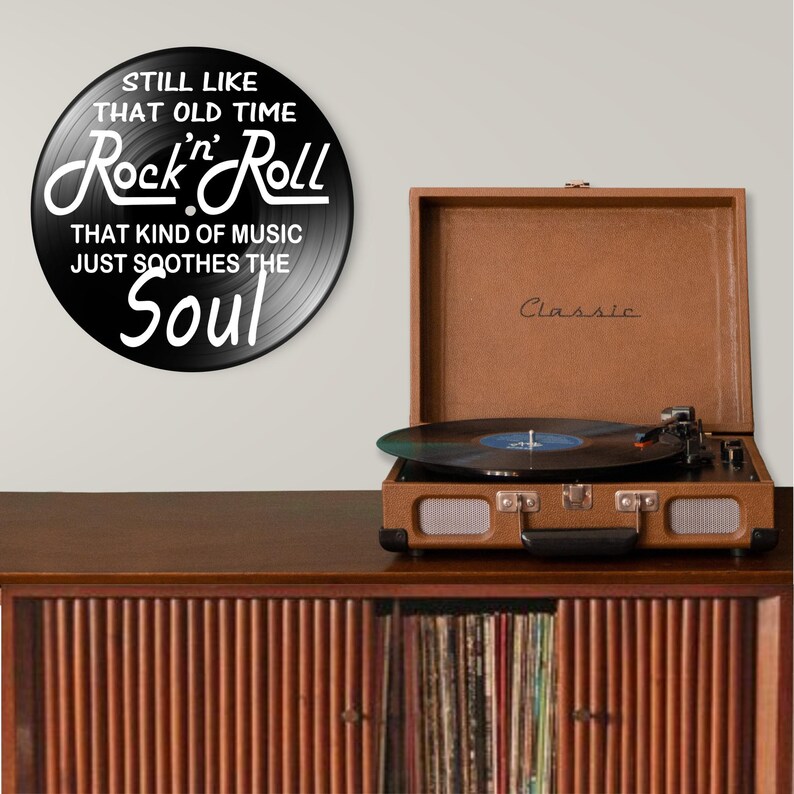 K&ouml;nnte beinhalten: Ein brauner Vintage-Plattenspieler mit einer Vinyl-Schallplatte, die auf einem Holzschrank abgespielt wird. Ein schwarzer runder Wandaufkleber mit den Worten "STILL LIKE THAT OLD TIME Rock 'n' Roll THAT KIND OF MUSIC JUST SOOTHES THE Soul" befindet sich an der Wand.