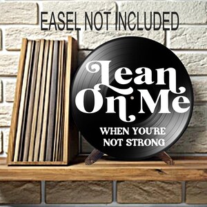 May include: A black vinyl record with the words "Lean On Me" in white script. The record is propped up on a wooden stand. The text "When You're Not Strong" is below the title. A wooden storage box is to the left.