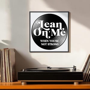 May include: A framed print featuring a black vinyl record design with the words "Lean On Me" in white, along with the phrase "When You're Not Strong." A black record player sits below the print, with stacks of vinyl records on either side.