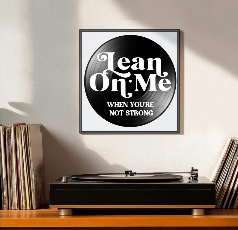 May include: A framed print featuring a black vinyl record design with the words "Lean On Me" in white, along with the phrase "When You're Not Strong." A black record player sits below the print, with stacks of vinyl records on either side.