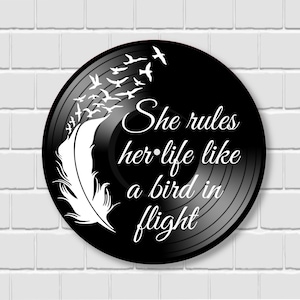 May include: A black vinyl record with white text that reads "She rules her life like a bird in flight." A white feather and flock of birds are also featured in the design. The record is set against a white brick wall.