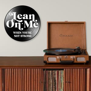 May include: A black vinyl record with the text "Lean On Me" and "When You're Not Strong" in white lettering. A brown vintage record player is on a wooden cabinet, with a record playing. The cabinet has a collection of records.