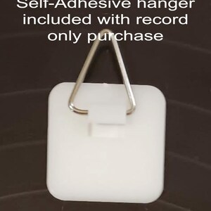 May include: A white, self-adhesive hanger with a metal triangle hook. The hanger is square with rounded corners. Text at the top reads "Self-Adhesive hanger included with record only purchase."