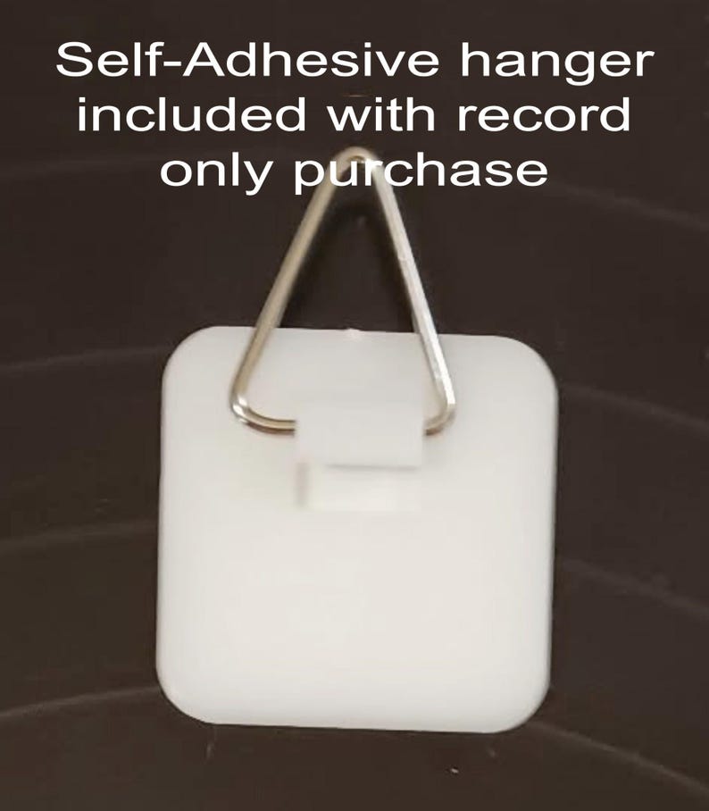 May include: A white, self-adhesive hanger with a metal triangle hook. The hanger is square with rounded corners. Text at the top reads "Self-Adhesive hanger included with record only purchase."