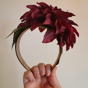 Puede incluir: Una diadema de flores de color burdeos con hojas verdes. La diadema está hecha de un material marrón y se sostiene en una mano.