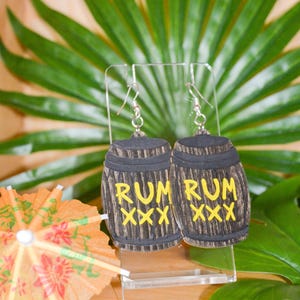 May include: A pair of novelty earrings shaped like wooden barrels. The earrings are dark brown with yellow text that reads "RUM XXX". The earrings are displayed on a clear stand with a tropical background.
