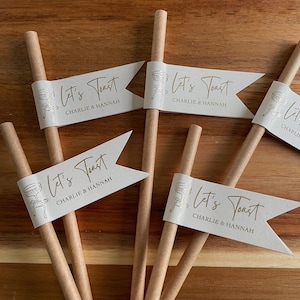 May include: Paper straws with white flag-shaped toppers. The flags read "Let's Toast" and "Charlie & Hannah" in gold script. The straws are light brown, and the flags have a simple illustration of a champagne glass.