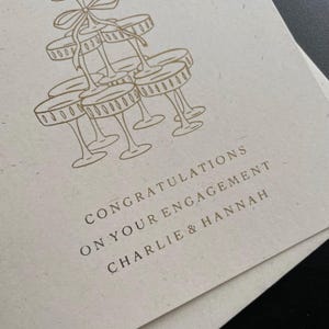 personalised your big news calls for a toast congratulations on your engagement, on ivory card with gold foil text with ivory envelope for engaged couple with tower of wine glasses.