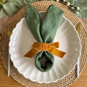 May include: An Easter table setting with a white, scalloped plate holding a green linen napkin shaped like bunny ears. A gold velvet ribbon secures the napkin. The plate sits on a woven placemat, with gold cutlery visible.