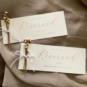 May include: Two ivory-colored wedding place cards with gold script that reads "Reserved". One card is for the "Father of the Groom" and the other for "Bridesmaid Hannah". Each card is decorated with a small bundle of dried flowers tied with white string.