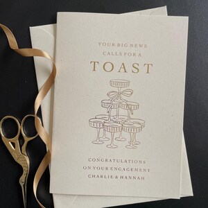 personalised your big news calls for a toast congratulations on your engagement, on ivory card with gold foil text with ivory envelope for engaged couple with tower of wine glasses.
