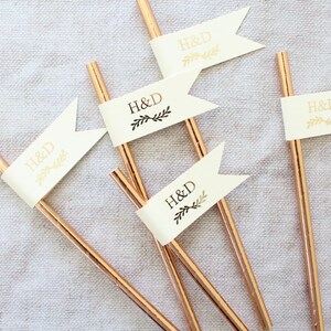 May include: Five gold straws with white flags attached. Each flag has the letters "H&D" printed in gold with a small black floral design.