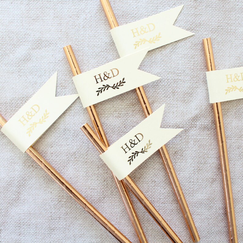 May include: Five gold straws with white flags attached. Each flag has the letters "H&D" printed in gold with a small black floral design.