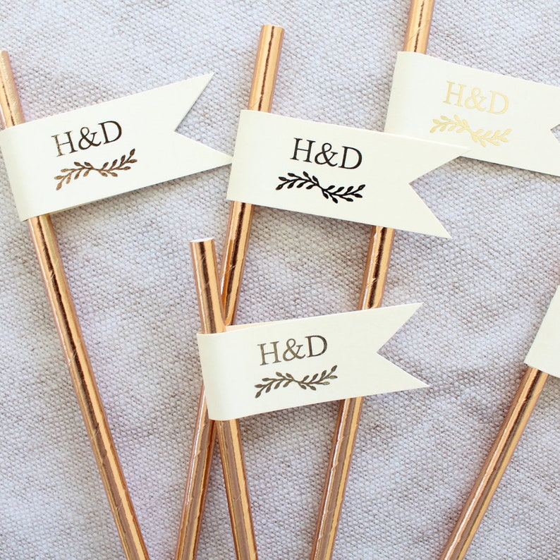 May include: Four gold straws with white flags attached. Each flag has the letters "H&D" printed in black with a small leaf design.
