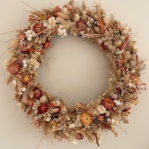 May include: A circular wreath composed of dried flowers and grasses in shades of burnt orange, deep red, cream, and beige. The arrangement features various textures including wheat stalks, fluffy bunny tails, seed pods, and small blossoms.