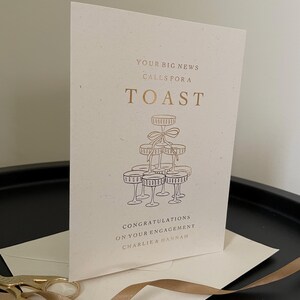 personalised your big news calls for a toast congratulations on your engagement, on ivory card with gold foil text with ivory envelope for engaged couple with tower of wine glasses.