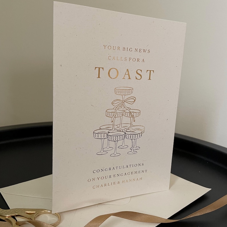 personalised your big news calls for a toast congratulations on your engagement, on ivory card with gold foil text with ivory envelope for engaged couple with tower of wine glasses.