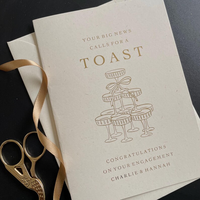 personalised your big news calls for a toast congratulations on your engagement, on ivory card with gold foil text with ivory envelope for engaged couple with tower of wine glasses.