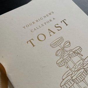 personalised your big news calls for a toast congratulations on your engagement, on ivory card with gold foil text with ivory envelope for engaged couple with tower of wine glasses.