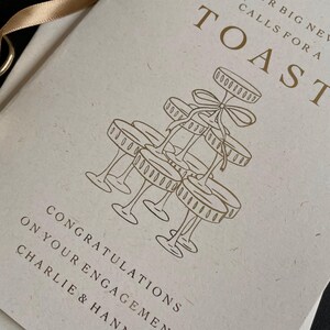 personalised your big news calls for a toast congratulations on your engagement, on ivory card with gold foil text with ivory envelope for engaged couple with tower of wine glasses.