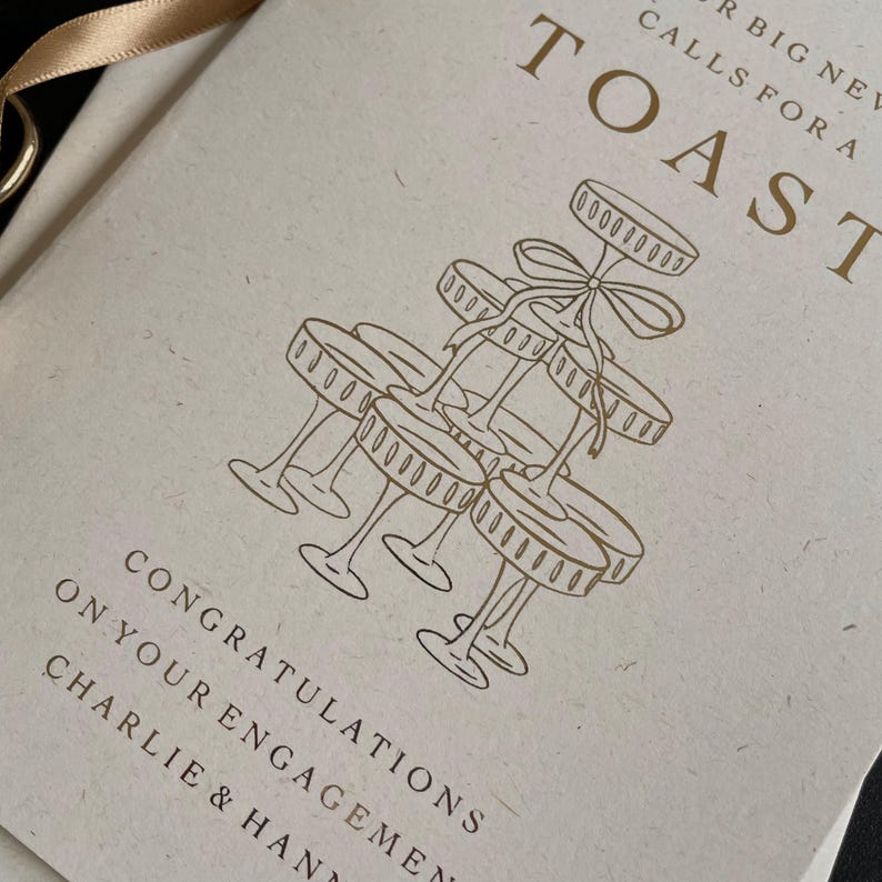 personalised your big news calls for a toast congratulations on your engagement, on ivory card with gold foil text with ivory envelope for engaged couple with tower of wine glasses.
