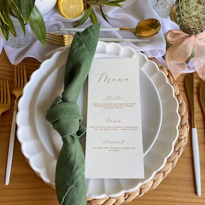 May include: A formal table setting with a white scalloped plate, gold utensils, and a green napkin. A menu card with gold lettering lists the starter, main course, and dessert. The table is set on a woven placemat.