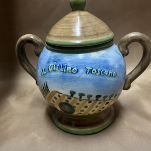 Puede incluir: Un azucarero de cerámica con tapa y dos asas. El cuenco está pintado con un cielo azul y un diseño de paisaje toscano. El texto "Il Villino Toscano" está escrito en el cuenco.