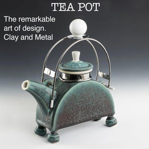 May include: A teal ceramic teapot with a silver lid and a silver metal handle. The teapot has a white knob on the lid and four small feet. The teapot is sitting on a white surface. The text "TEA POT" is written in black letters above the teapot. The text "The remarkable art of design. Clay and Metal" is written in black letters below the teapot.