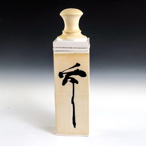 May include: A rectangular, cream-colored ceramic container with a black, stylized Japanese character. The container has a light beige lid and a light beige, rounded stopper. The container is approximately 8 inches tall.