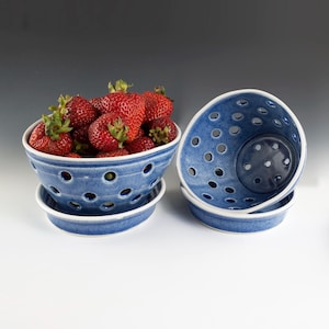 May include: Blue ceramic berry bowls with a white rim and numerous circular perforations. One bowl is filled with fresh, ripe strawberries. The bowls are stacked on matching saucers. The design is ideal for serving fresh fruit.
