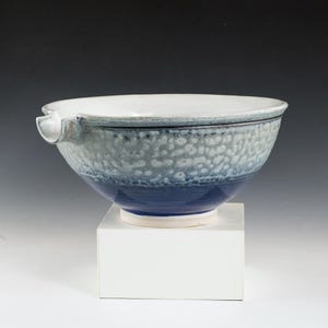 May include: A ceramic bowl with a spout, featuring a white interior and a textured exterior with shades of blue and gray. The bowl sits on a white square pedestal, showcasing its unique design and craftsmanship. The bowl's dimensions are not visible.