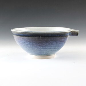 Peut inclure: Un bol en céramique fait à la main avec un glaçage bleu et blanc. Le bol a une forme arrondie avec une petite poignée et un intérieur blanc. L'extérieur présente des nuances de bleu et de gris, créant un effet de dégradé. Le bol repose sur une base blanche.