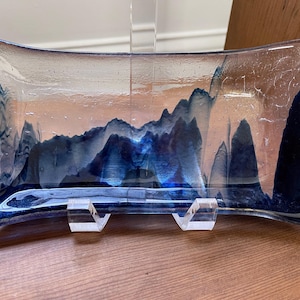 May include: A rectangular glass dish with a blue and white abstract design resembling mountains. The dish is supported by two clear acrylic stands. The glass has a slightly curved shape and is displayed on a wooden surface.