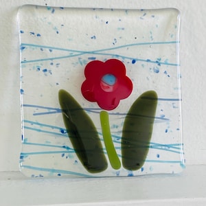 May include: A square glass art piece featuring a red flower with a blue center, green stem, and two green leaves. The background is clear with blue lines and dots, creating a floral design.