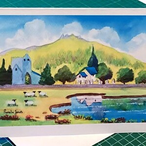May include: A watercolor painting of a blue church with a white roof, a white house with a blue roof, and a small pond with a blue reflection. The scene is set in a green valley with a yellow hill in the background. There are sheep in the field and the artist's signature, "Pamela Jones", is in the bottom left corner.