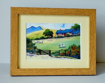 May include: Watercolor painting of a rural landscape with a farmhouse, sheep, and a gate. The artwork is framed in a light brown wooden frame with a cream-colored mat. The painting features green fields, blue mountains, and a blue sky.