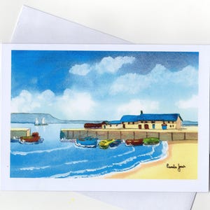 May include: Watercolor art print of a coastal scene featuring a harbor with boats, a building, and a beach under a blue sky with clouds. The artwork is on a white card with a white envelope.