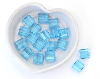 May include: A white heart-shaped dish filled with light blue glass beads. The beads are square shaped and have a slightly frosted finish. Some beads are scattered outside the dish.