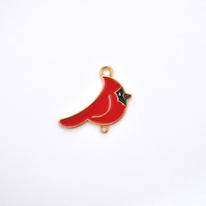 May include: A gold-toned metal charm shaped like a cardinal bird with bright red enamel filling the body and a black enamel patch around the beak. The charm features two small circular loops, one at the top and one at the bottom for attachment.