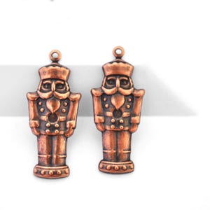 May include: Two copper-colored metal nutcracker charms. Each charm is a miniature figure of a nutcracker soldier, complete with a crown, mustache, and uniform. The charms have a loop at the top for attaching to jewelry or other crafts.