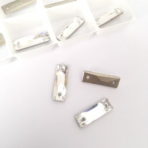 May include: A close-up of clear crystal rhinestones in a rectangular shape with two holes. The rhinestones are arranged on a white surface.