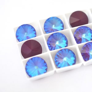 May include: A white plastic tray with compartments filled with blue and purple rhinestones. The rhinestones are round and have a faceted design.