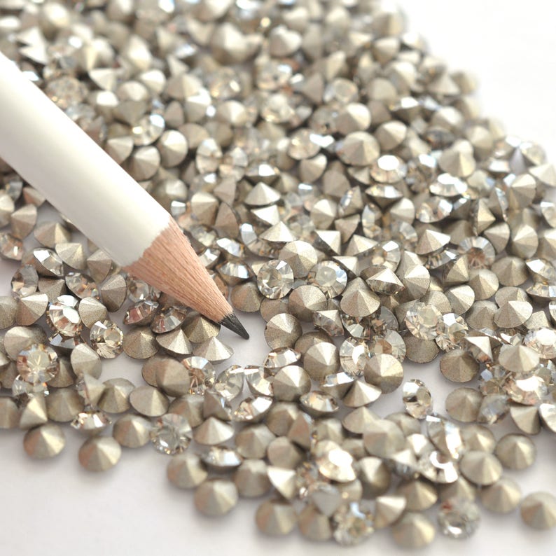 May include: A white pencil lies on a white surface surrounded by a large quantity of small, silver, faceted rhinestones.