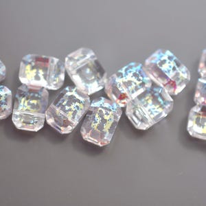 May include: A collection of clear, rectangular beads with iridescent, multicolored patterns. The beads are arranged on a gray surface, showcasing their multifaceted design and light refraction.
